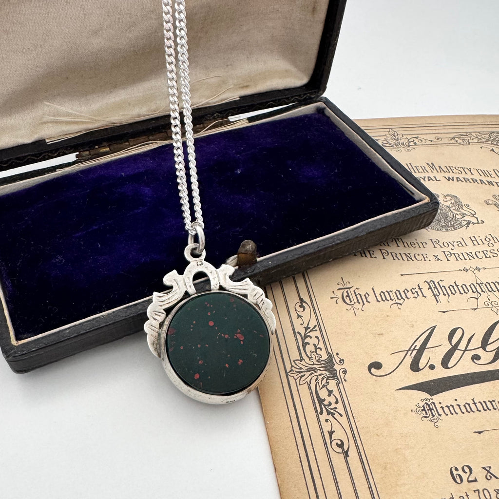 Silver 1905 antique watch fob necklace with a bloodstone insert in an open jewelry box on an old document.