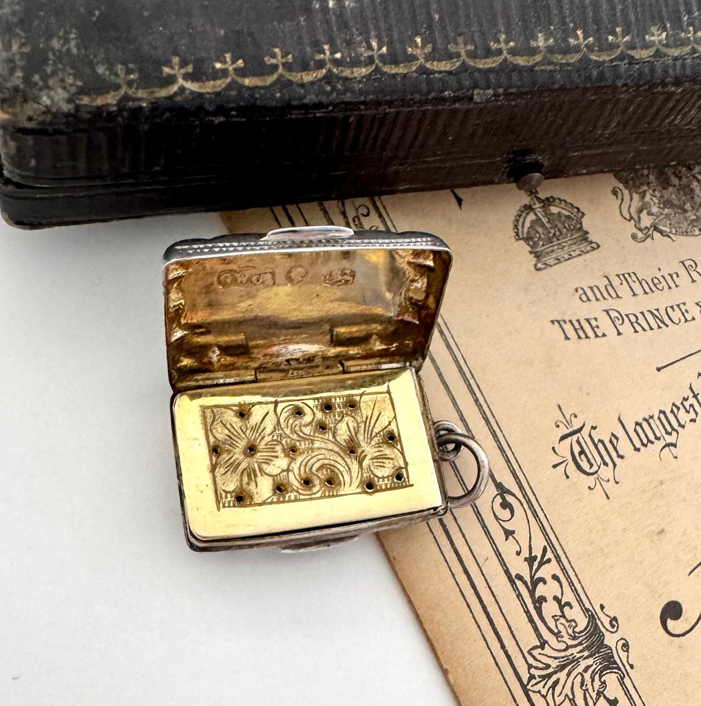 Antique 1864 sterling silver vinaigrette on an antique advertising material for a photography studio and a vintage jewellery box. The vinaigrette is open showing the grill inside.
