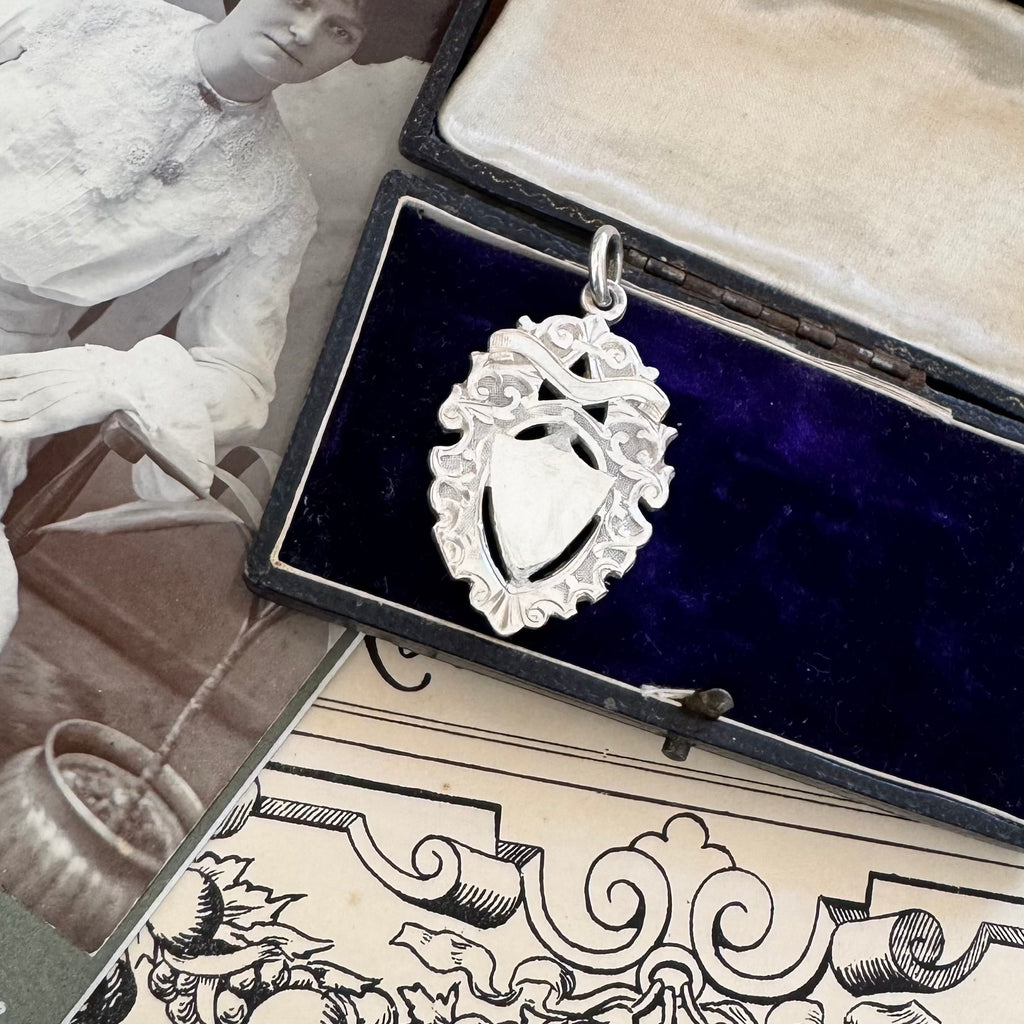 Antique silver watch fob pendant in an open jewelry box with a vintage-style illustration of a woman and decorative elements.