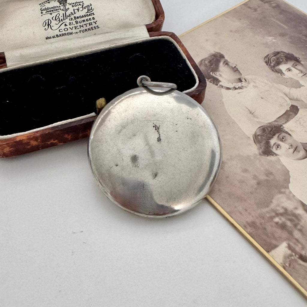 An antique Adie & Lovekin round silver toned compact pendant with a small loop at the top, shown resting on a white surface beside an open vintage presentation box and an old sepia-toned portrait photograph.