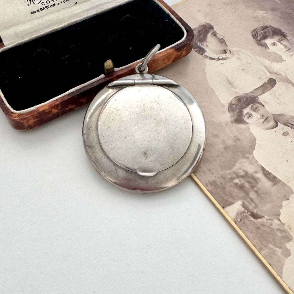 An antique silver oval compact mirror with a hinged lid, displayed next to its open brown leatherette case and an old sepia-toned photograph.