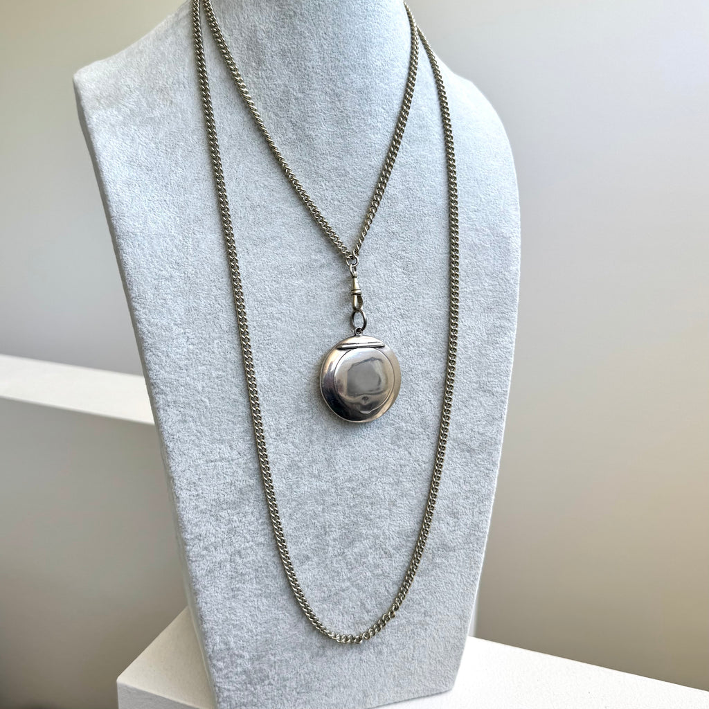 A silver-toned guard chain necklace with a round antique compact locket pendant, displayed on a light gray fabric jewelry bust.