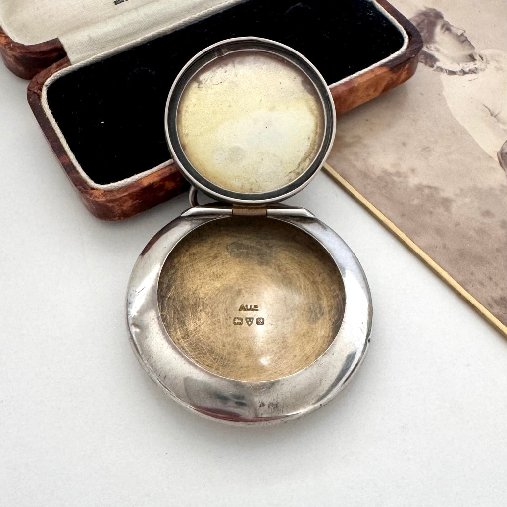 An antique Adie & Lovekin open round silver compact or powder case with a hinged lid, shown on a light surface beside its dark velvet-lined wooden box and an old printed page. The inside of the lid has a pale cream-colored insert or cover.