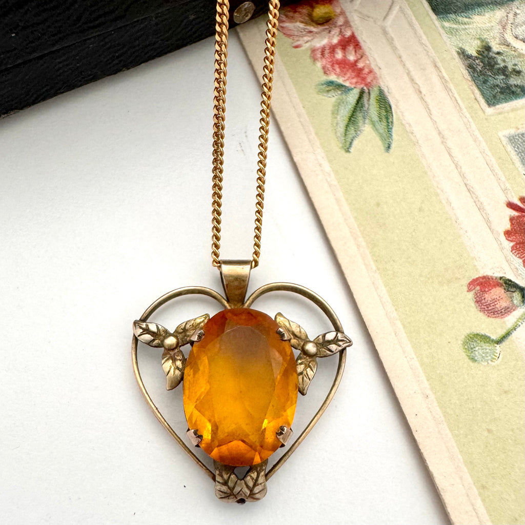 Antique heart-shaped pendant with amber-colored stone on a gold chain, placed on a textured surface.