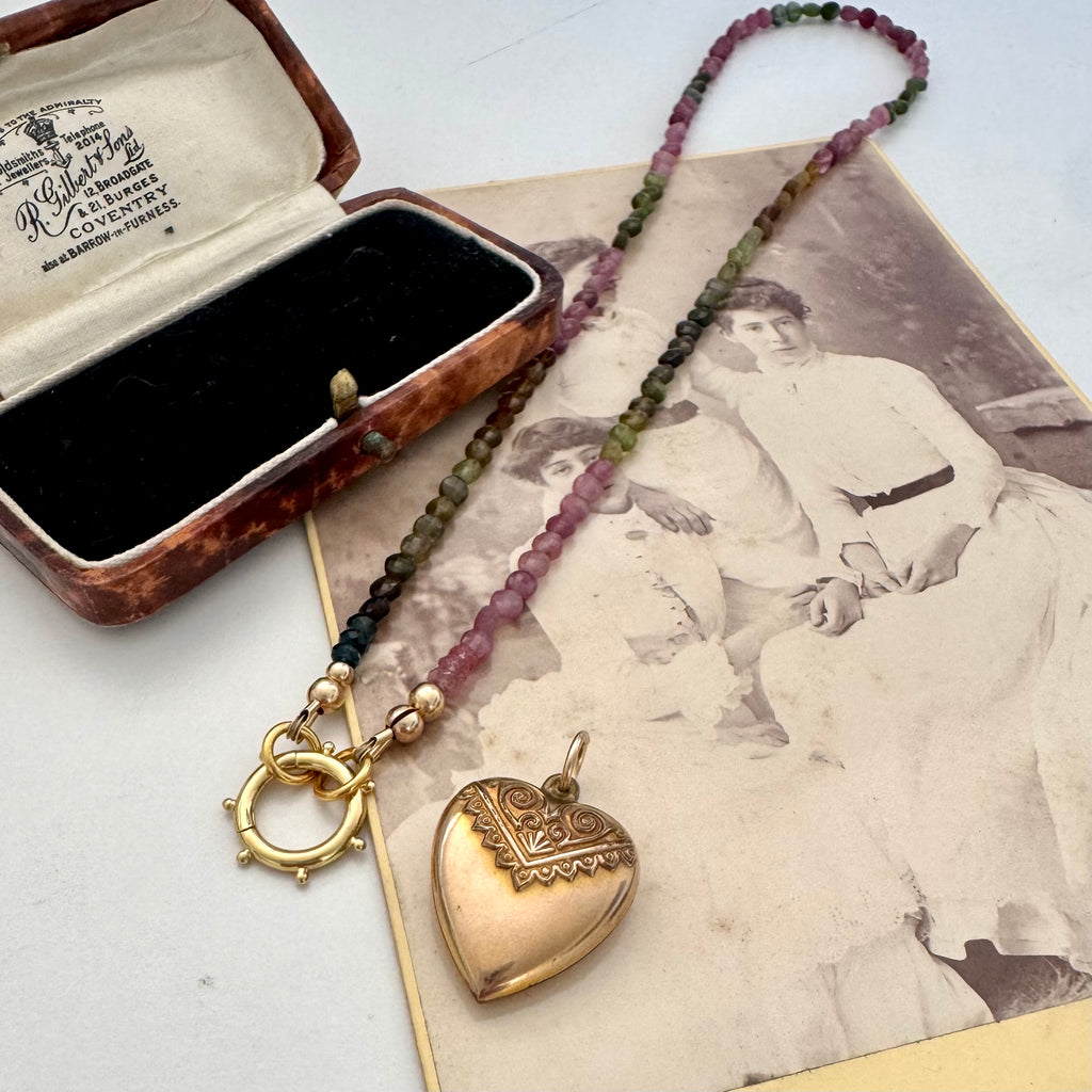 A beaded tourmaline necklace with an antique Etruscan revival 9ct gold lined heart pendant, displayed on a vintage-style card next to an open jewelry box. The beads transition from dark green to pinkish-purple, and the pendant is gold-toned with engraved details.
