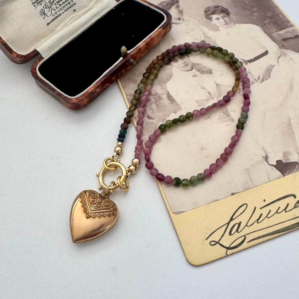 A beaded tourmaline necklace with an antique Etruscan revival 9ct gold lined heart pendant, displayed on a vintage-style card next to an open jewelry box. The beads transition from dark green to pinkish-purple, and the pendant is gold-toned with engraved details.