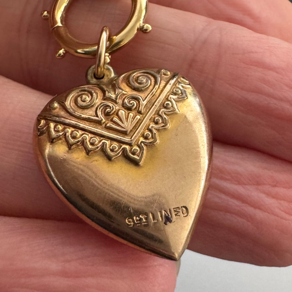 A close-up of an antique gold heart-shaped pendant held in a hand. The pendant features an engraved top section with scrollwork and a decorative border, and the word 9ct lined is stamped on the lower right side.