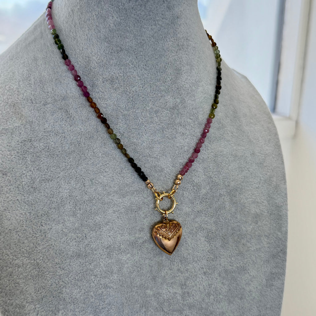 A beaded necklace with a heart-shaped pendant displayed on a gray fabric mannequin bust. The necklace features small faceted beads in pink, green, and black, connected by gold-toned metal links, with a gold heart pendant hanging from a ring clasp.