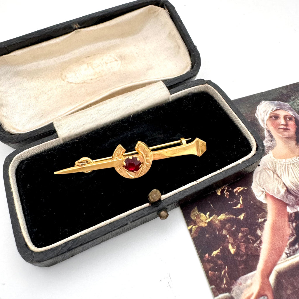 Vintage 9ct gold lined horseshoe and nail brooch with a red gemstone on a white surface with a blurred background