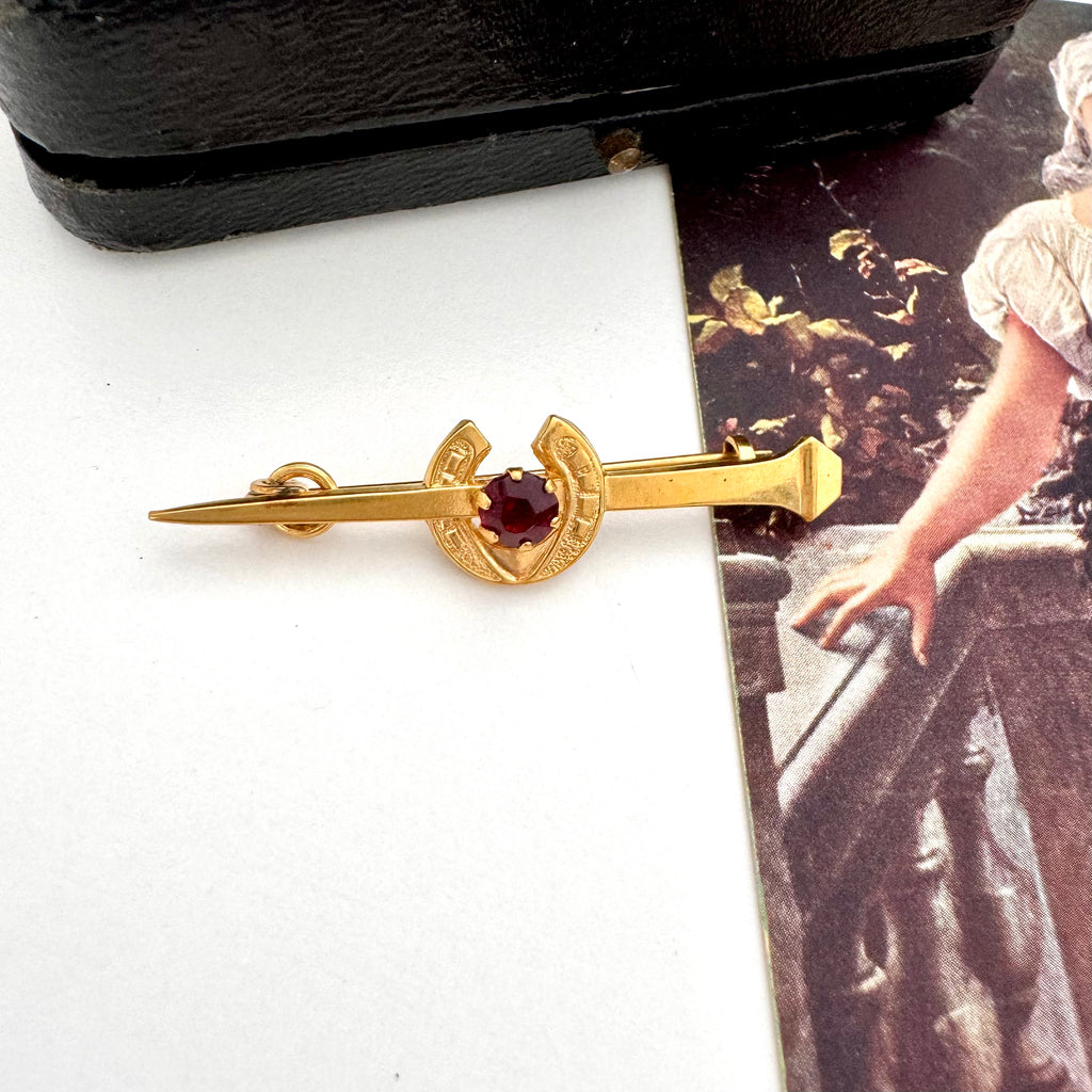 Vintage 9ct gold lined horseshoe and nail brooch with a red gemstone on a white surface with a blurred background
