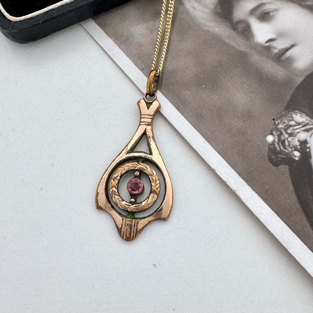 A vintage-style pendant necklace with a gold-toned chain and a teardrop-shaped pendant in rose gold and silver, featuring a central pink gemstone. The pendant is displayed on a light gray surface next to a black box corner and a black-and-white vintage photograph.
