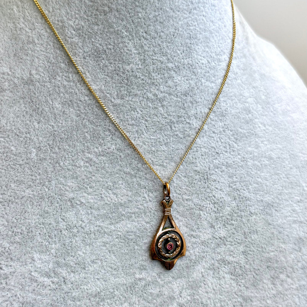 A gold chain necklace with a teardrop-shaped pendant featuring a central circular design with a red center, displayed on a textured light gray mannequin bust.