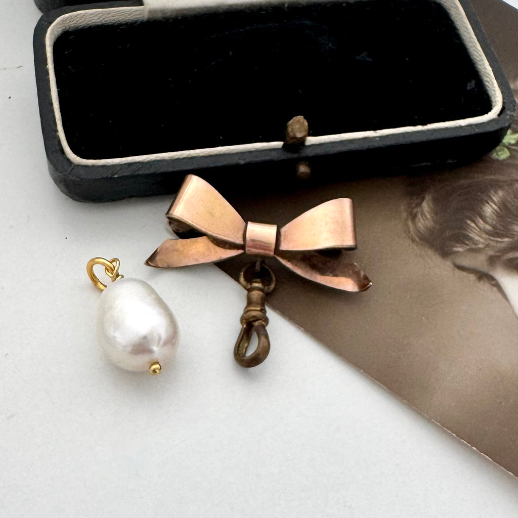 An Edwardian rolled rose gold bow-shaped brooch with a dangling clasp and a white baroque pearl pendant on a gold loop, displayed next to an open black jewelry box on a white surface.