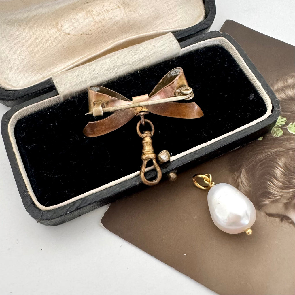 Back of Edwardian rolled gold bow brooch in warm metallic tones displayed inside an open black jewelry box, with a white teardrop pearl pendant on a brown card beside it.