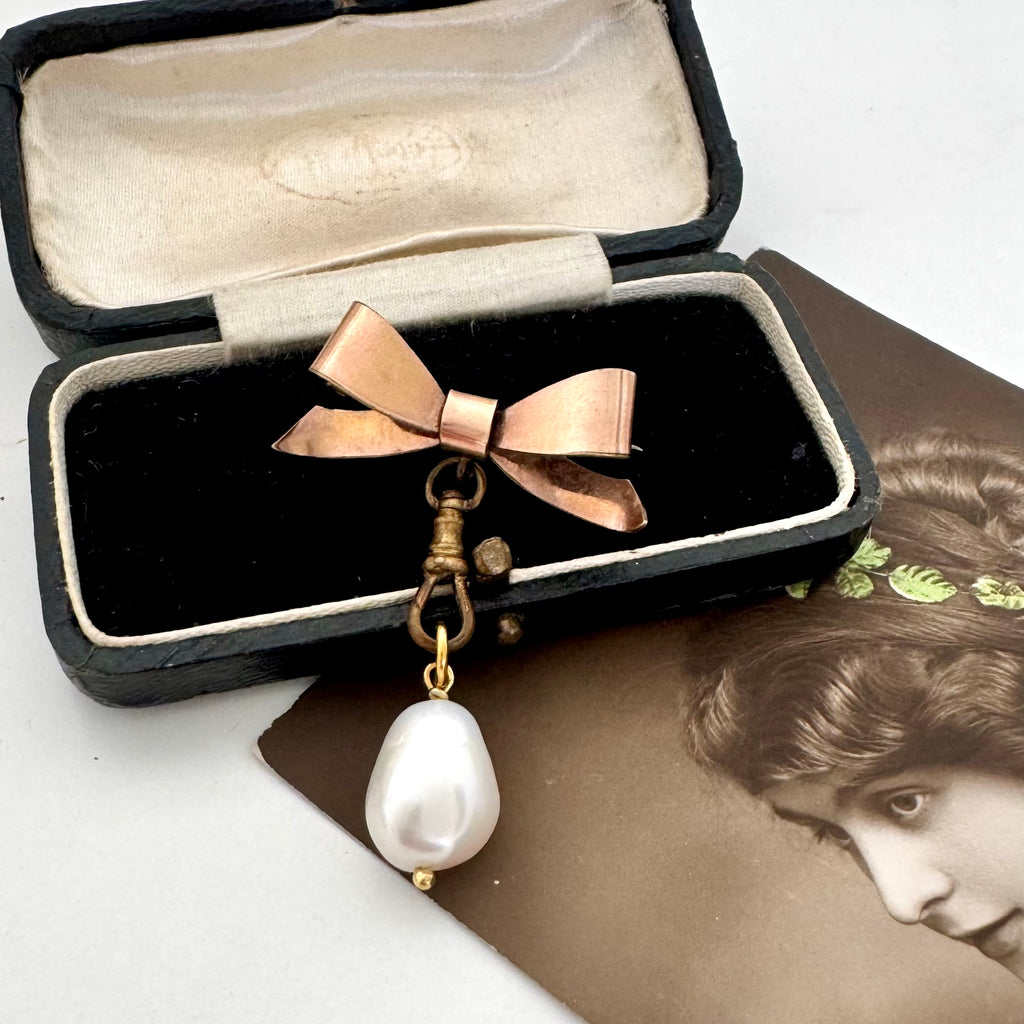 An Edwardian rolled gold bow brooch, gold-toned clasp, and a dangling white baroque pearl, displayed inside an open black presentation box with a cream velvet lining.