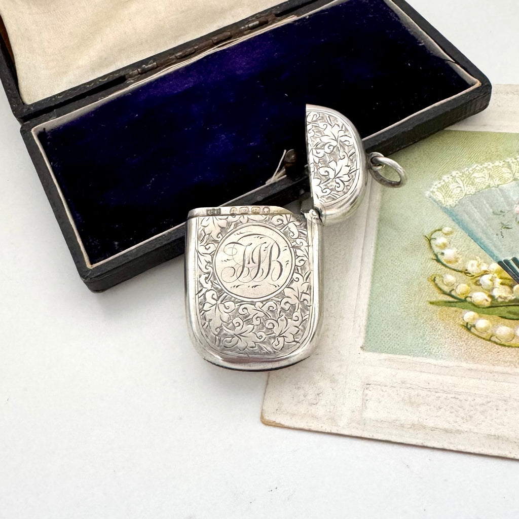 Antique sterling silver 1887 vesta case with intricate patterns on a white surface next to an open jewelry box and vintage card.