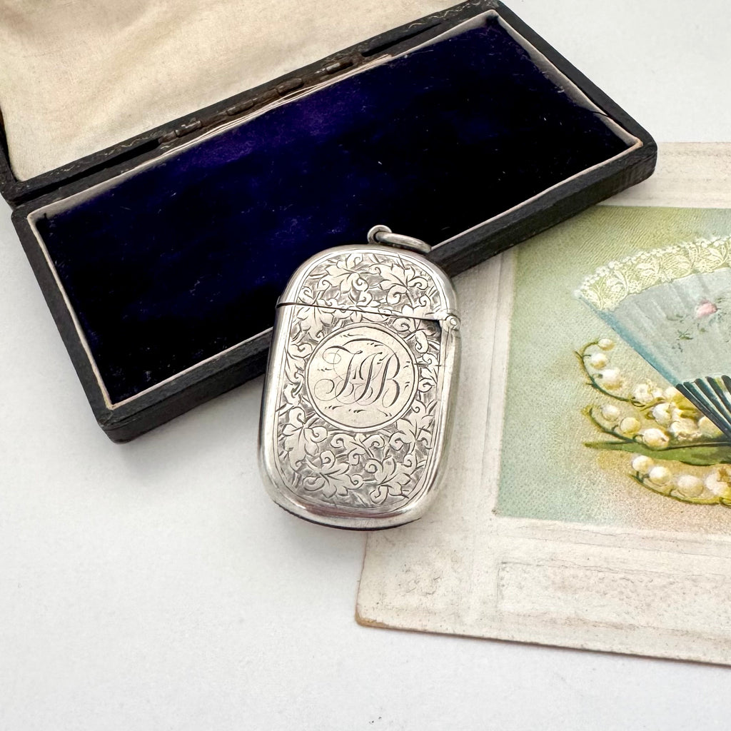 Antique sterling silver 1887 vesta case with intricate patterns on a white surface next to an open jewelry box and vintage card.