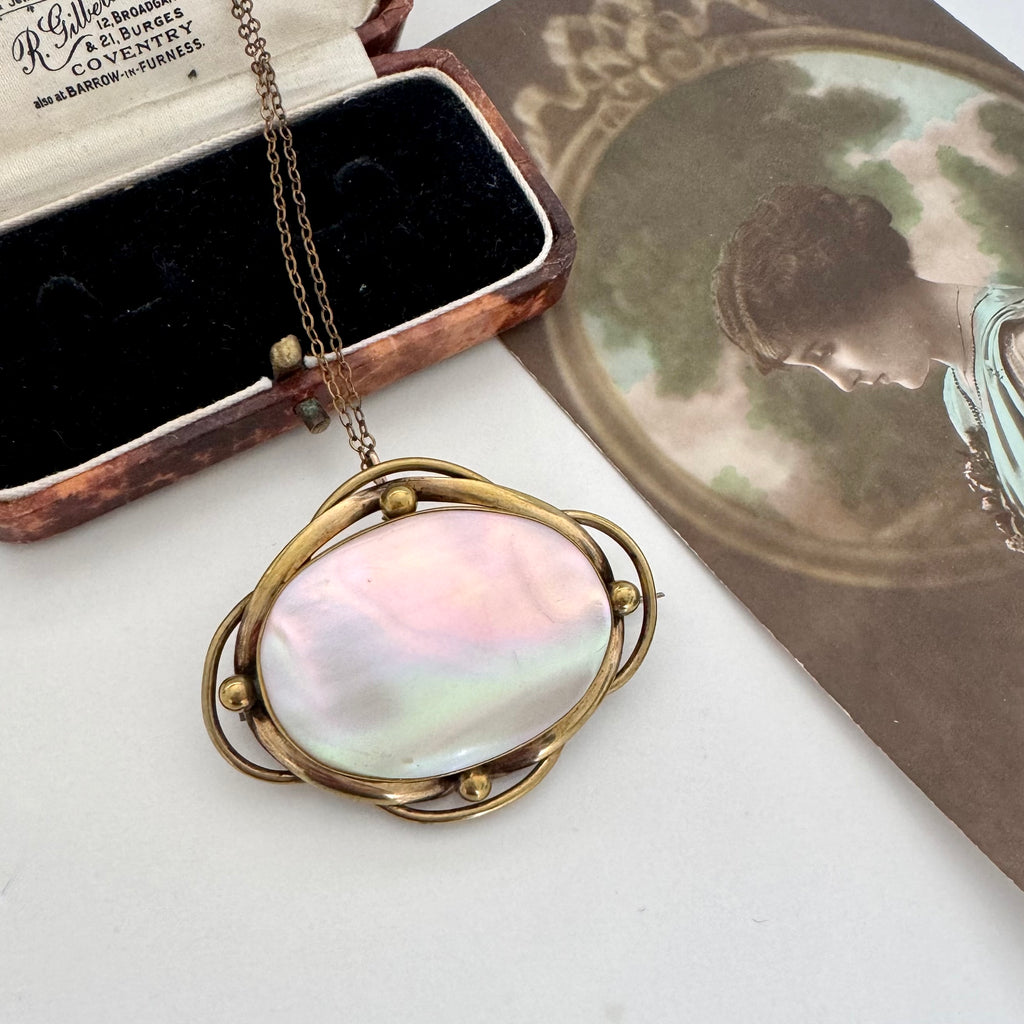 A Victorian oval brooch/pendant necklace with a iridescent mother-of-pearl center and an organic-shaped gold frame, displayed next to a open vintage presentation box and a decorative card.