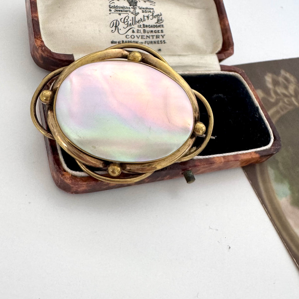 An antique oval mother-of-pearl brooch in a gold-toned setting, displayed inside its vintage wooden presentation box with a black velvet interior. The box lid features a printed paper insert with the brand 'R.