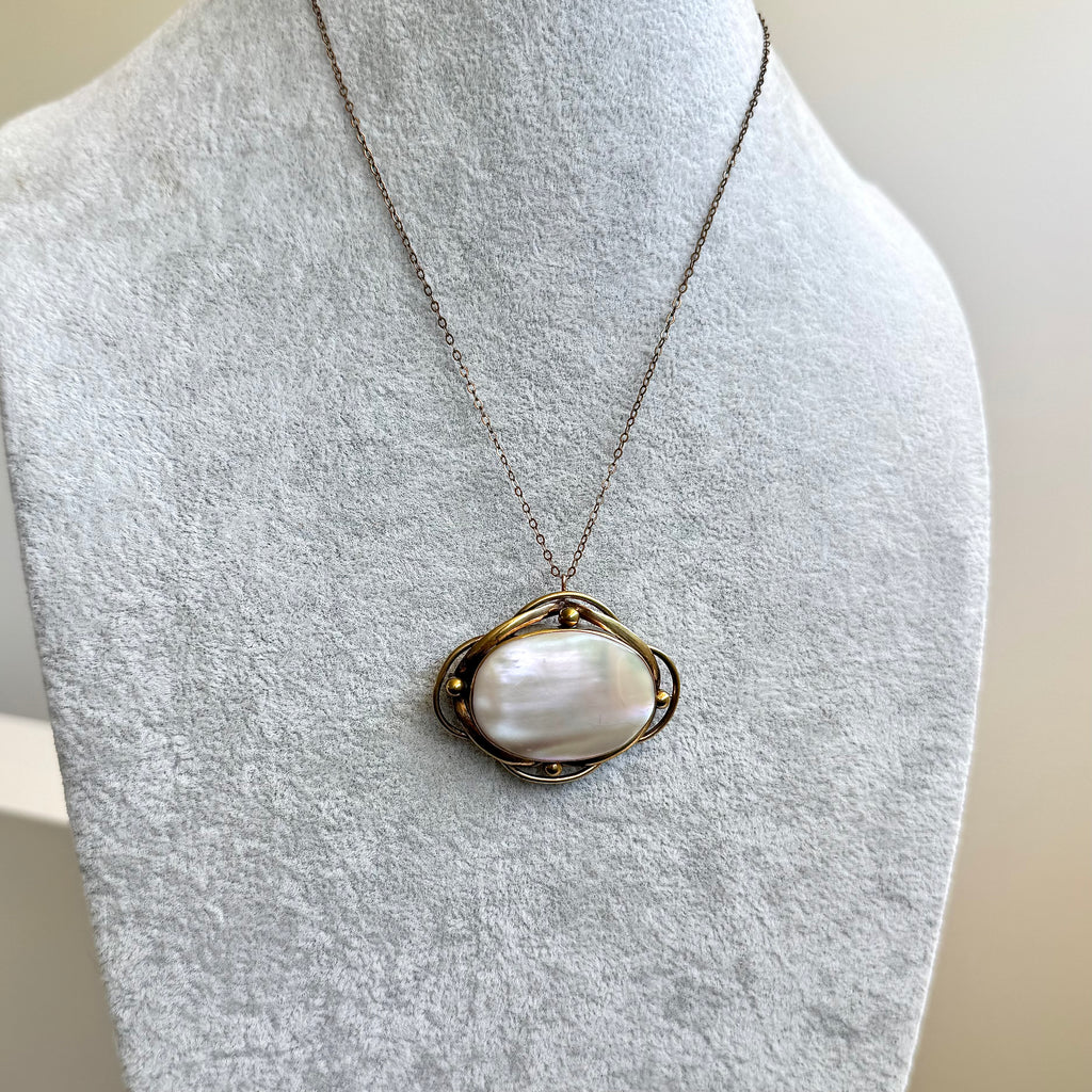 A delicate chain necklace with an antique oval pendant featuring a white mother-of-pearl center and a gold-toned metal border, displayed on a light gray fabric jewelry bust.