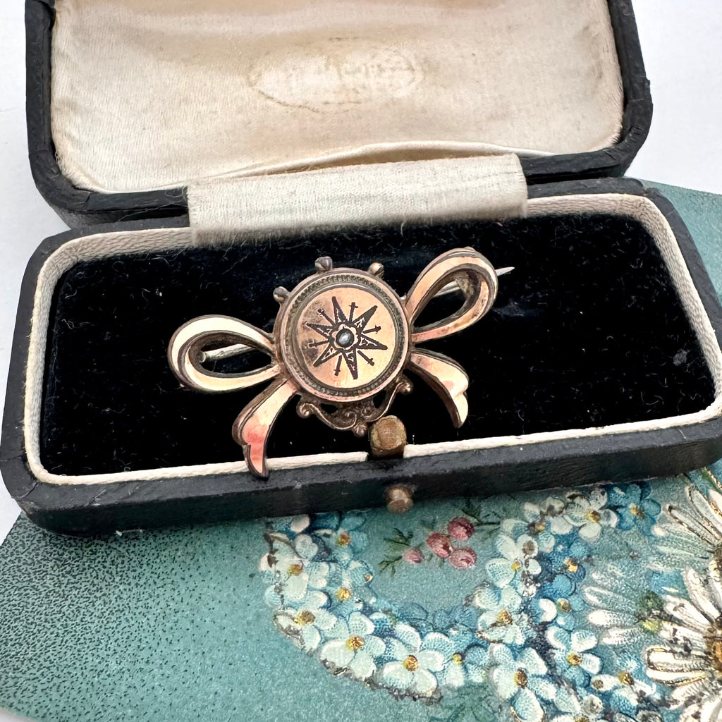 Antique Victorian star burst and bow-shaped brooch displayed on a vintage jewellery box and with a floral card in the background.