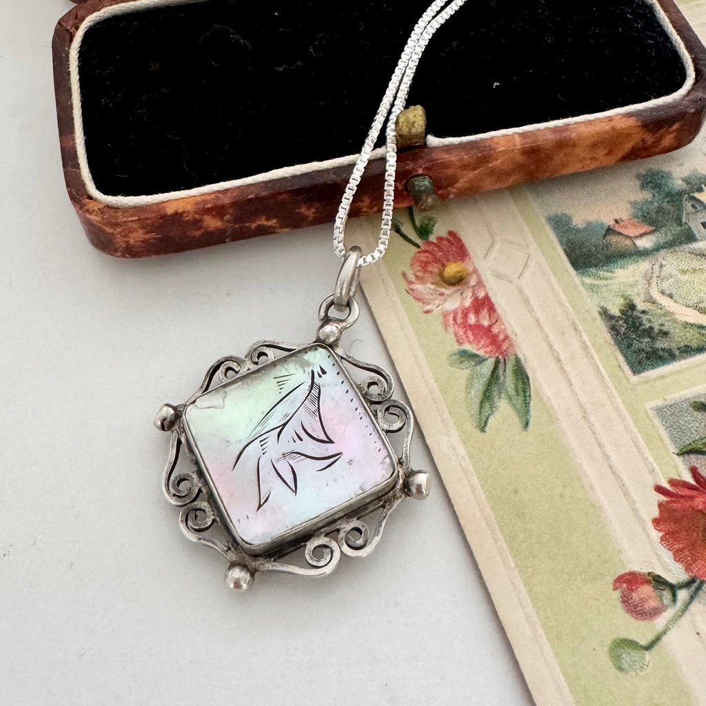 A silver-toned pendant necklace with a square iridescent shell inlay featuring a black ink sketch of a bird, displayed next to an open vintage jewelry box and a floral card.