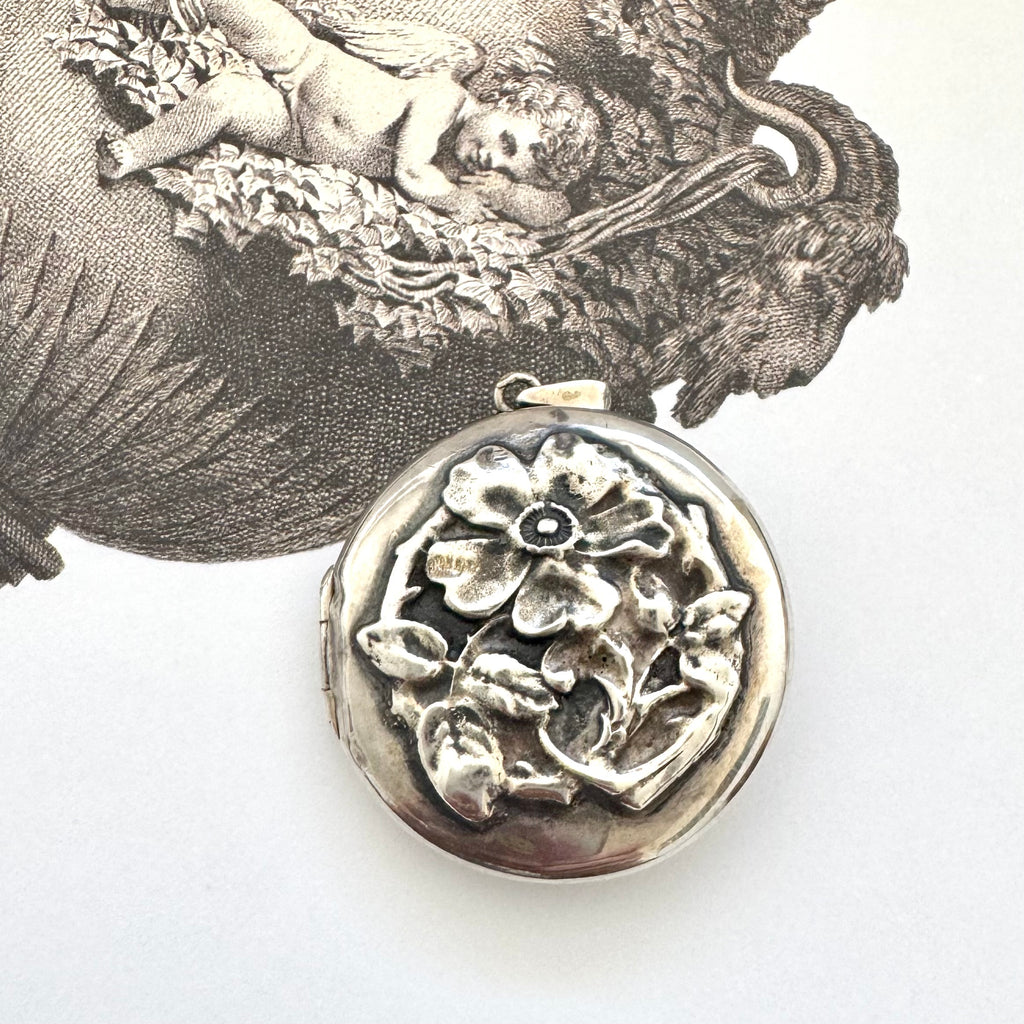Sterling silver art nouveau repousse floral locket on a white background with an engraved cherub design in the background.