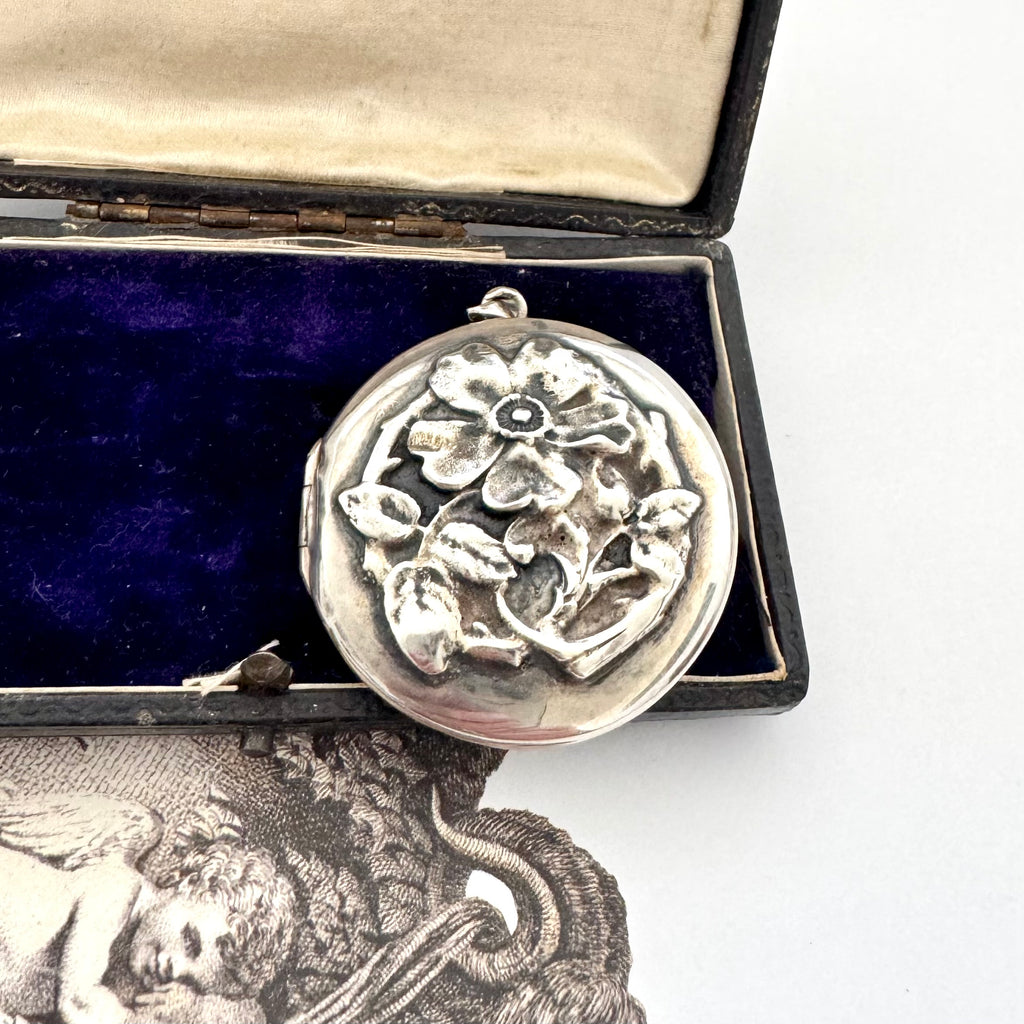 Sterling silver art nouveau repousse floral locket on a white background with an engraved cherub design in the background and on a box