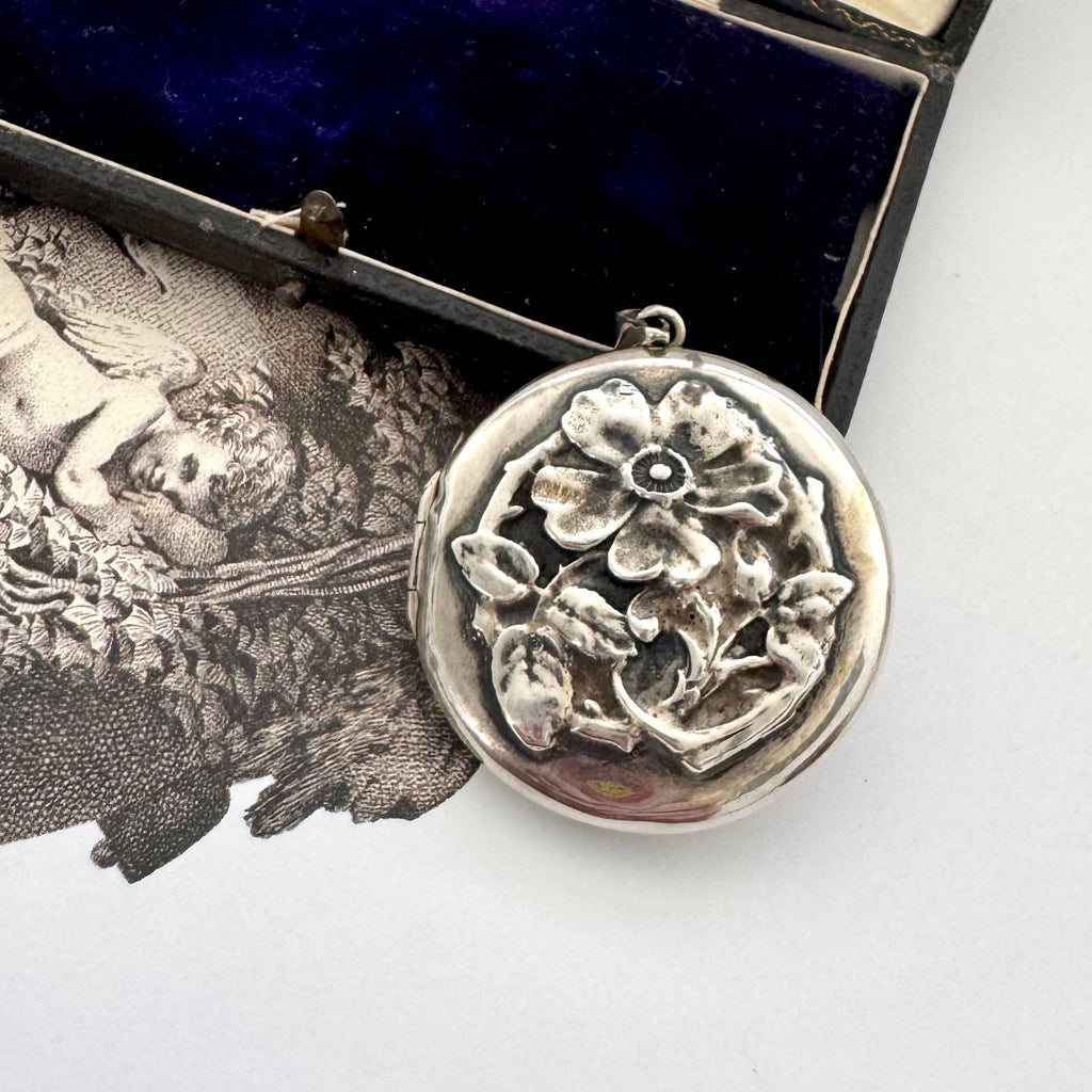 Sterling silver art nouveau repousse floral locket on a white background with an engraved cherub design in the background.