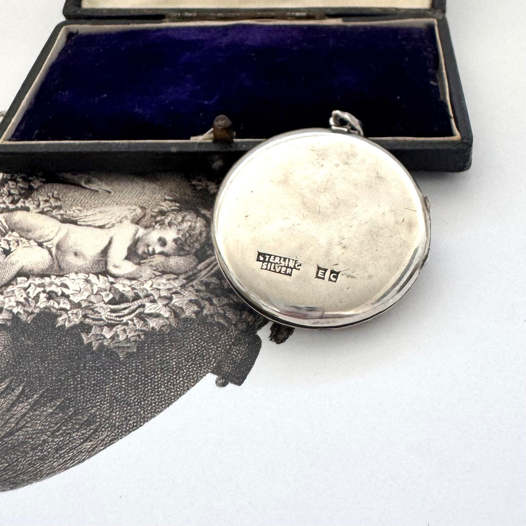 Back of sterling silver art nouveau repousse floral locket on a white background with an engraved cherub design in the background.