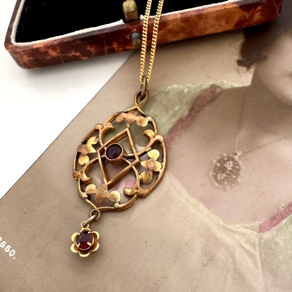 Antique Edwardian art nouveau lavalier necklace with a geometric openwork design and a small dangling flower charm, both set with dark red gemstones, displayed on a vintage illustrated card.