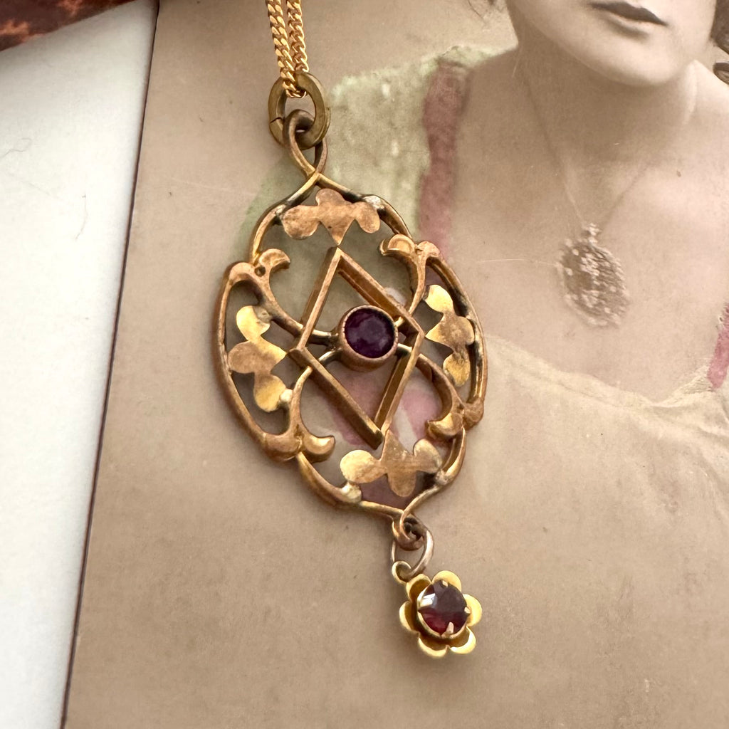 Antique Edwardian art nouveau lavalier necklace with filigree detailing, featuring a central purple gemstone and a dangling flower-shaped red gemstone, displayed against a vintage sepia-toned portrait background.