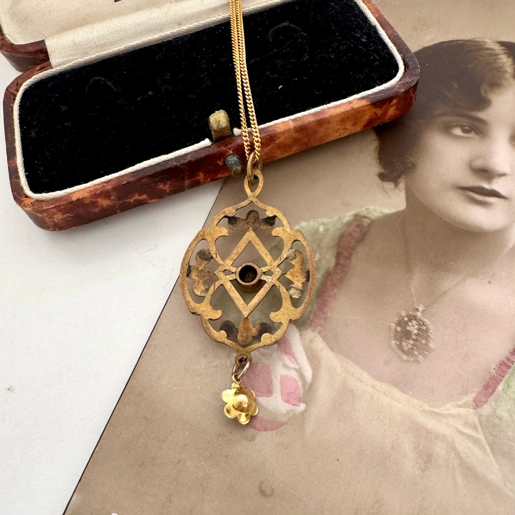 Back of antique Edwardian art nouveau lavalier pendant necklace with a decorative filigree design and a small dangling charm, displayed on top of an open vintage jewelry box over a sepia-toned portrait photo.