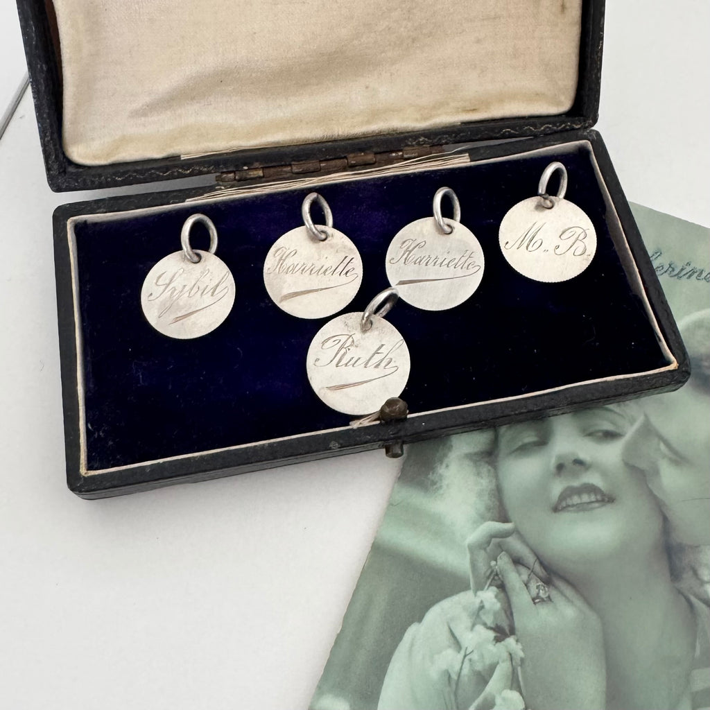 Set of silver charms with engraved names in an open jewelry box, with a vintage photo in the background.