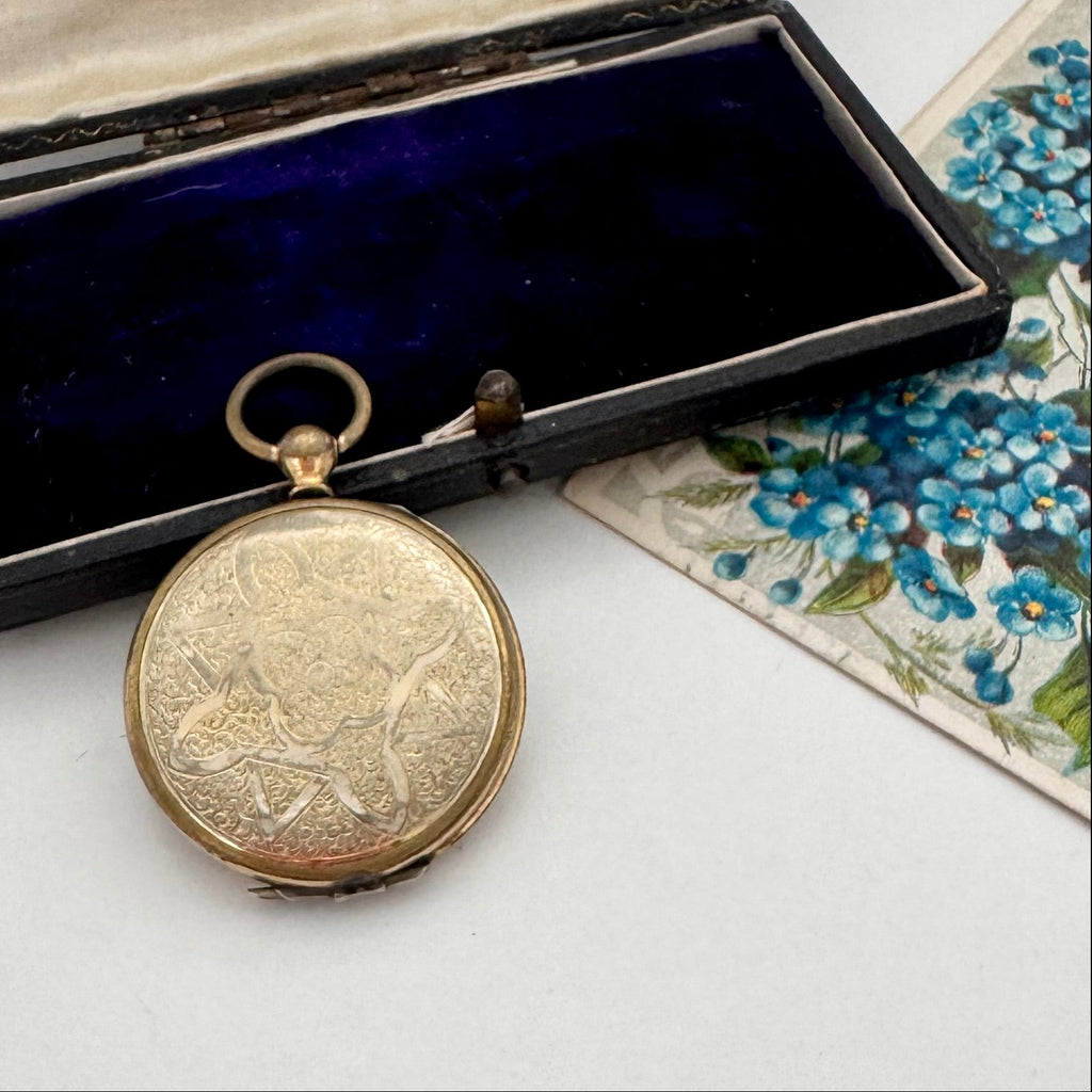 Antique pocket watch style locket with intricate design on a white surface next to an open jewelry box.