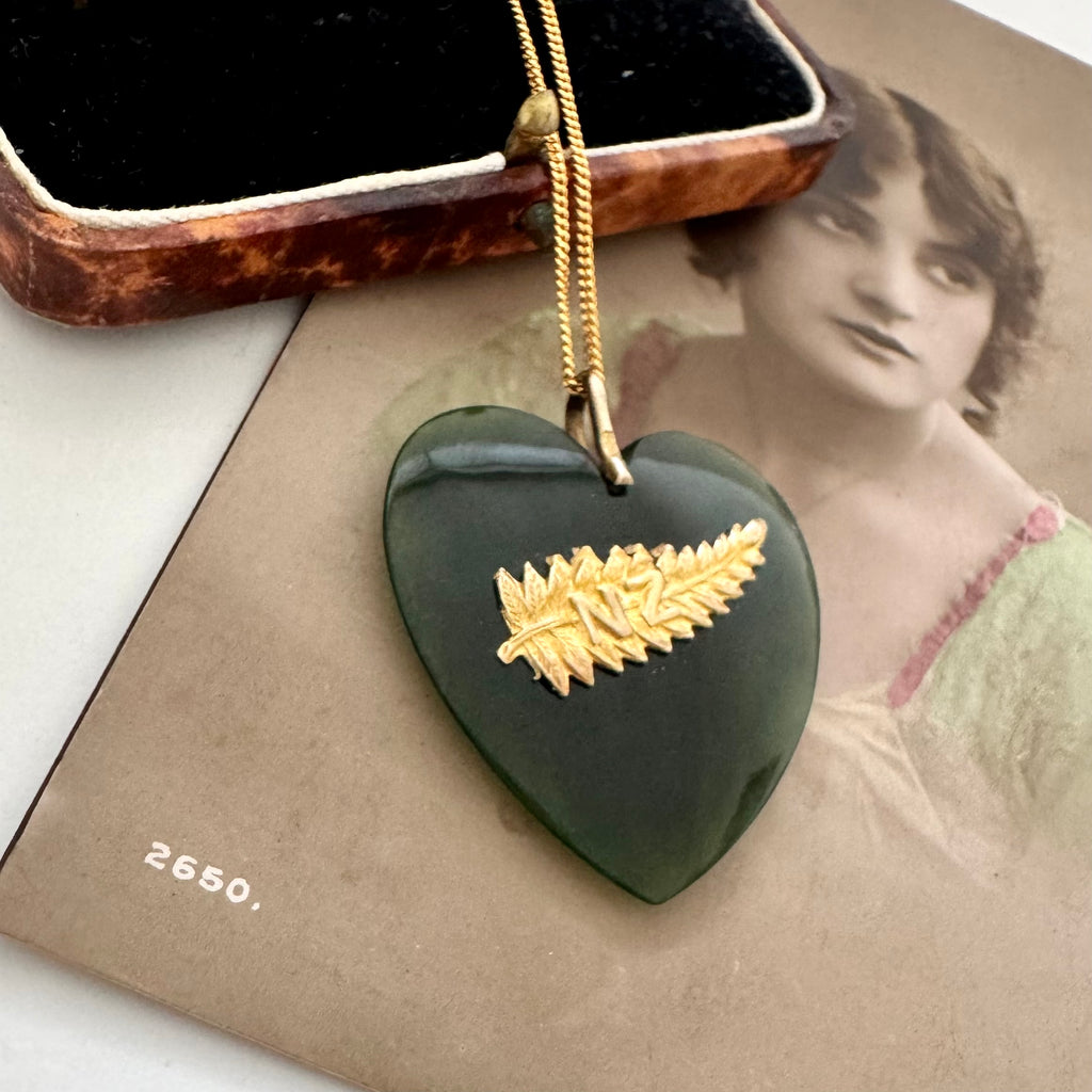 An antique heart-shaped dark green jade pounamu pendant necklace with a gold fern leaf inlay, resting on a vintage sepia-toned card next to its wooden presentation box.
