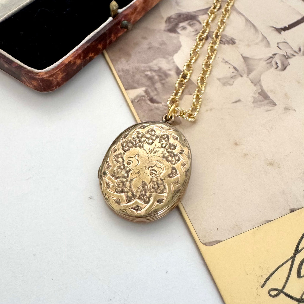 An antique oval gold locket pendant necklace with intricate floral engravings, resting on a vintage-style printed card next to an open jewelry box.