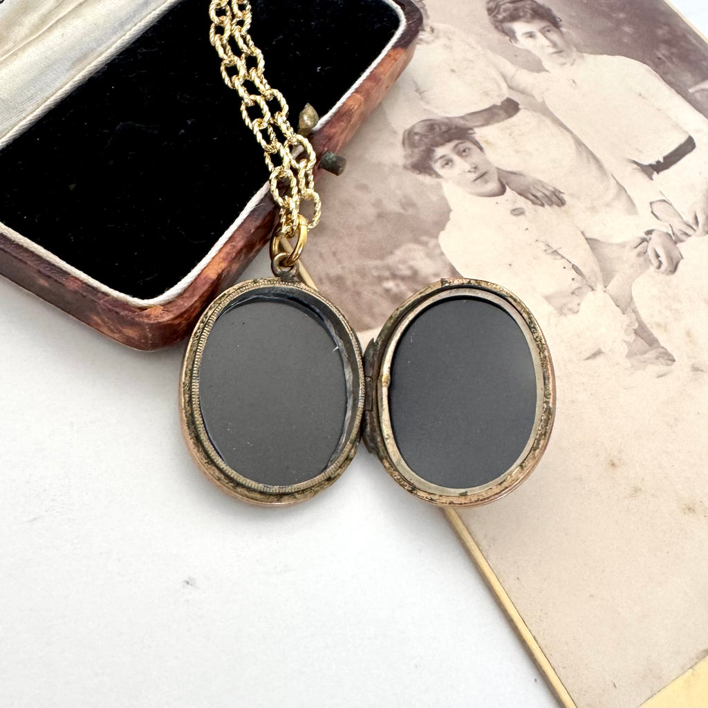 An antique oval locket necklace with dark glass panels and a gold-toned chain, displayed on a vintage photo print next to an open black velvet jewelry box.