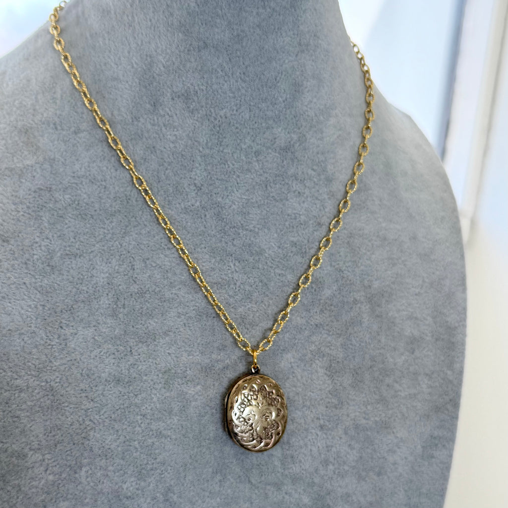 A gold chain necklace with an oval, textured pendant displayed on a gray velvet jewelry bust.