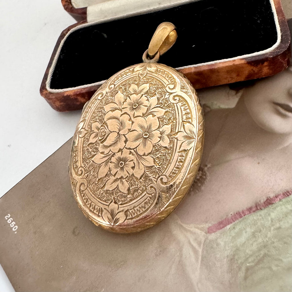 A Victorian oval gold pendant with intricate floral engravings, displayed next to its vintage brown velvet jewelry box on a white surface.