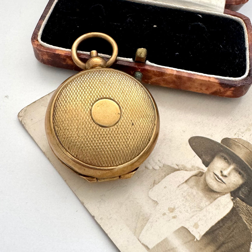 An antique rolled gold sovereign holder with a textured circular cover, displayed next to its open wooden presentation box with black velvet lining, with a vintage portrait photo in the background.