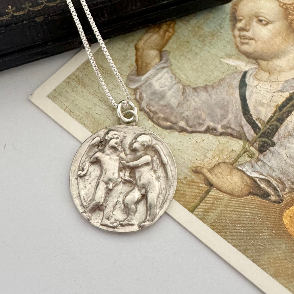 Antique silver pendant necklace on a gray surface with an old book and illustration in the background. The pendant features Cupid and Psyche in relief and has been converted from an antique button.