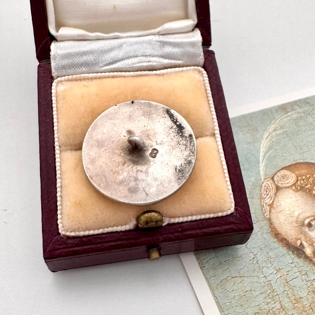 Antique silver button with relief sculpture in a wooden box on a white surface.