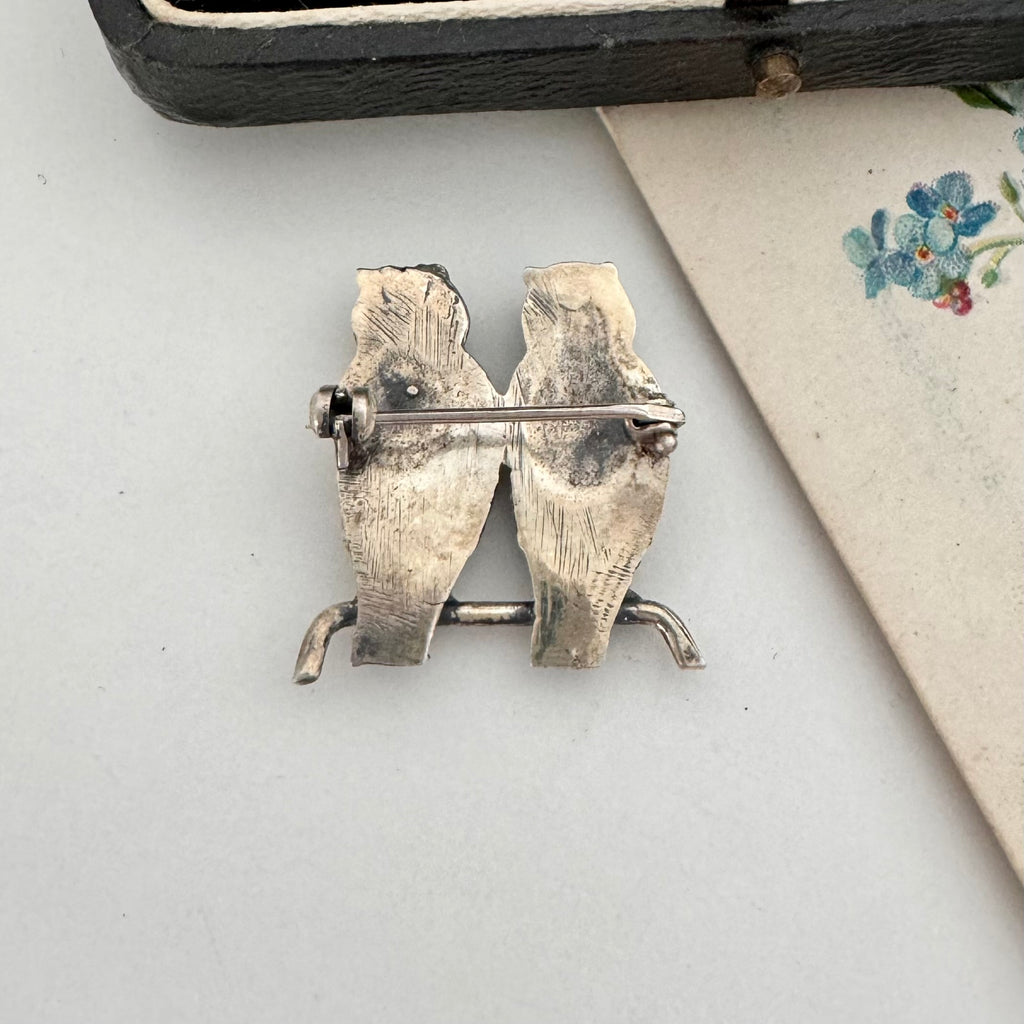 Silver bird-shaped brooch on a light gray surface with a floral-patterned fabric in the background.