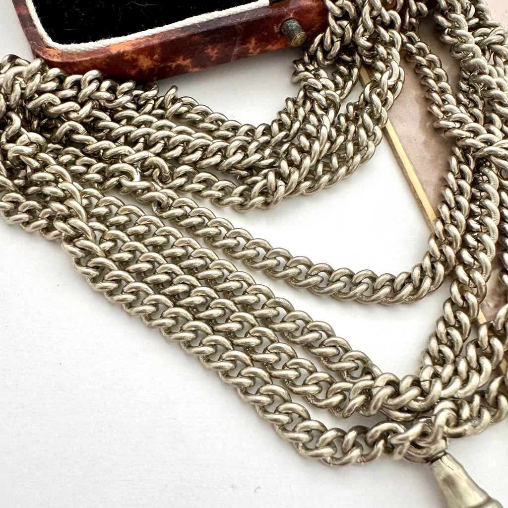 A close-up of a silver plated guard chain necklace draped across a white surface, with part of a brown marbled object visible in the upper left corner.