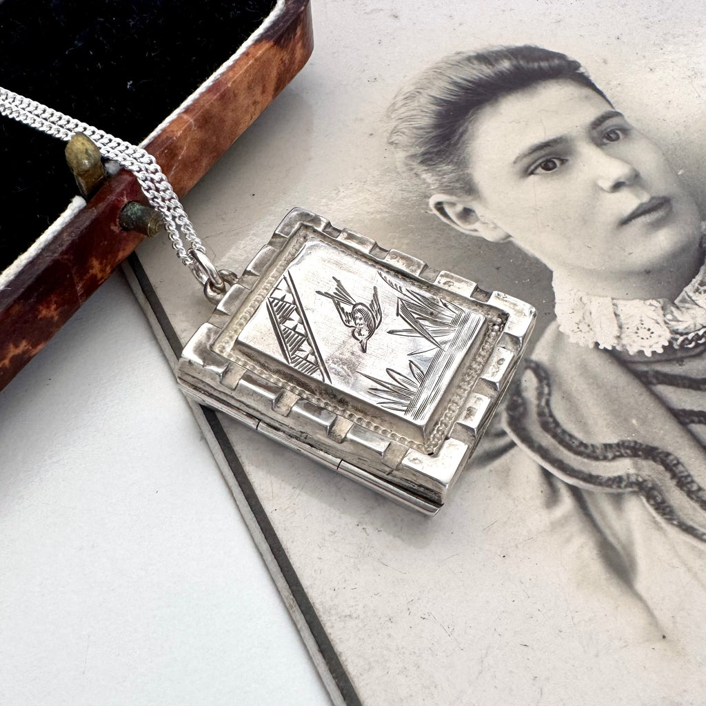 A silver locket pendant with engraved floral and bird motifs, resting on an antique black-and-white portrait print next to an open vintage jewelry box.