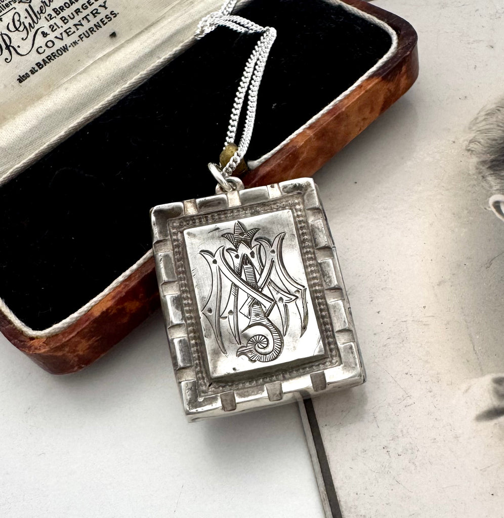 A silver pendant necklace with an engraved monogram for Auspice Mary, resting on its brown velvet-lined presentation box. The box lid shows the brand name 'Reginald & Barbour Coventry'.