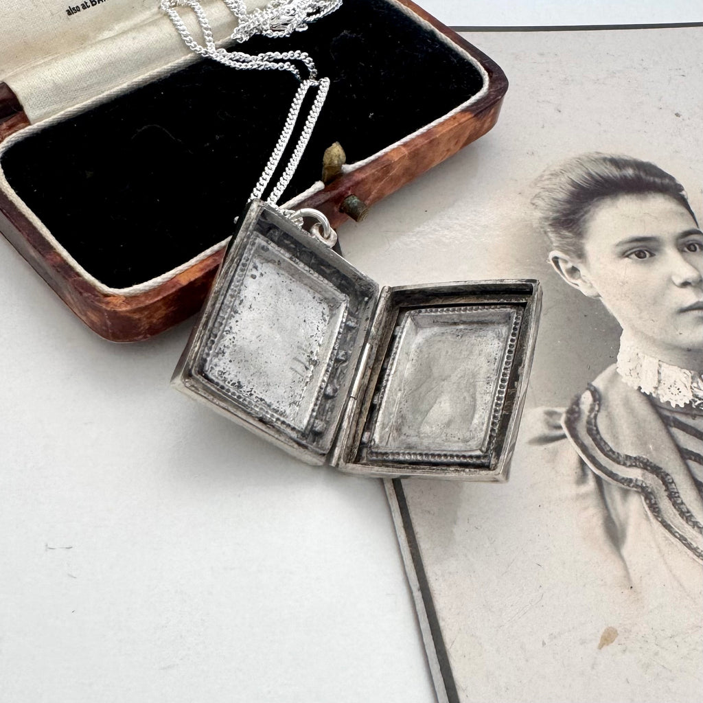 An open silver locket pendant necklace displayed on a white surface next to its vintage wooden presentation box and a sepia-toned portrait print.