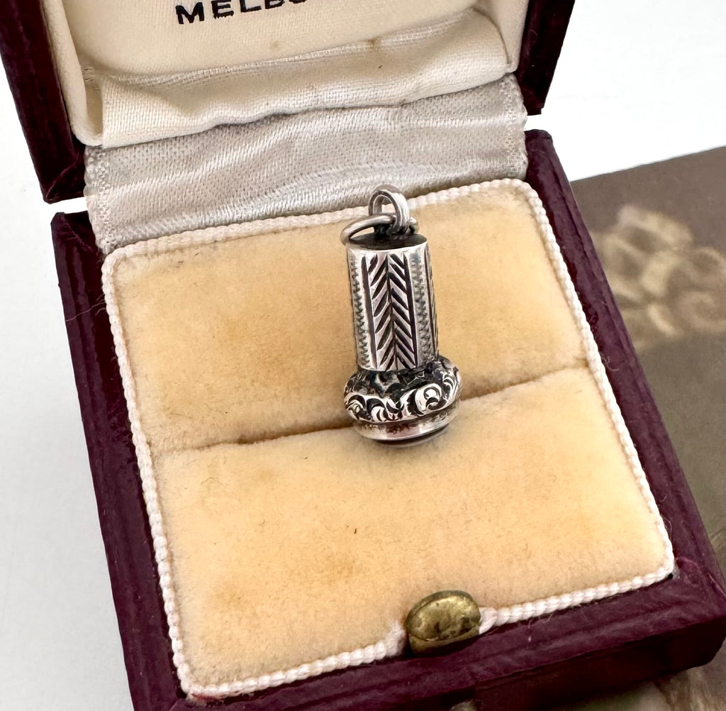 An antique miniature silver wax seal fob with engraved leaf-like detailing and a round pale blue stone, displayed inside an open burgundy jewelry box with cream velvet lining.
