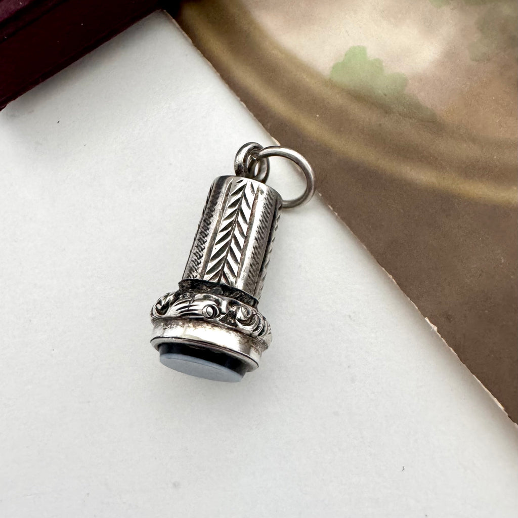 An antique silver miniature wax seal fob charm with engraved leaf and scroll patterns, resting on a white surface with a brown textured background.